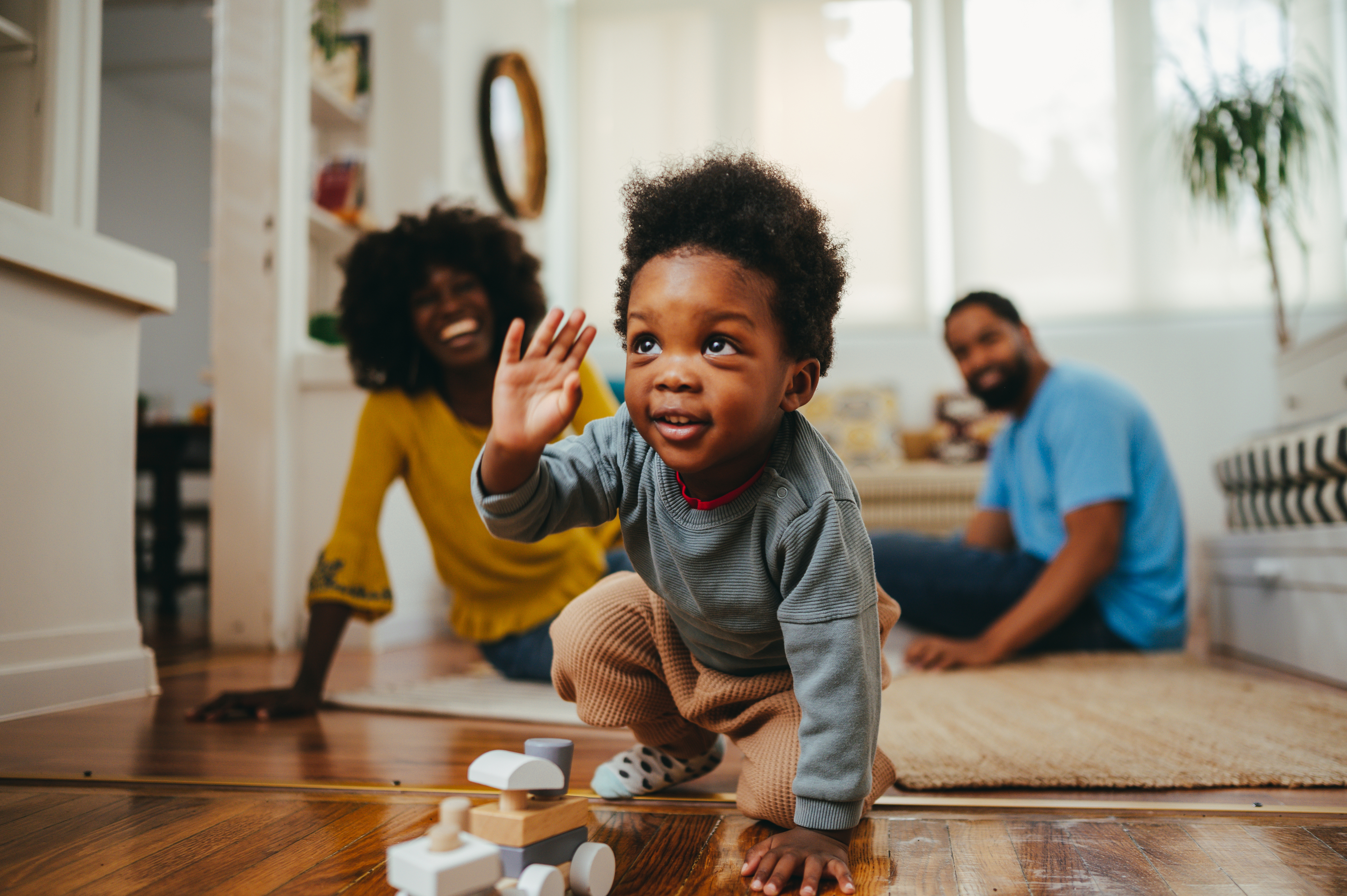 Parents play with their child who is crawling on the floor, wearing a gray shirt and light pants, with toy blocks in the foreground