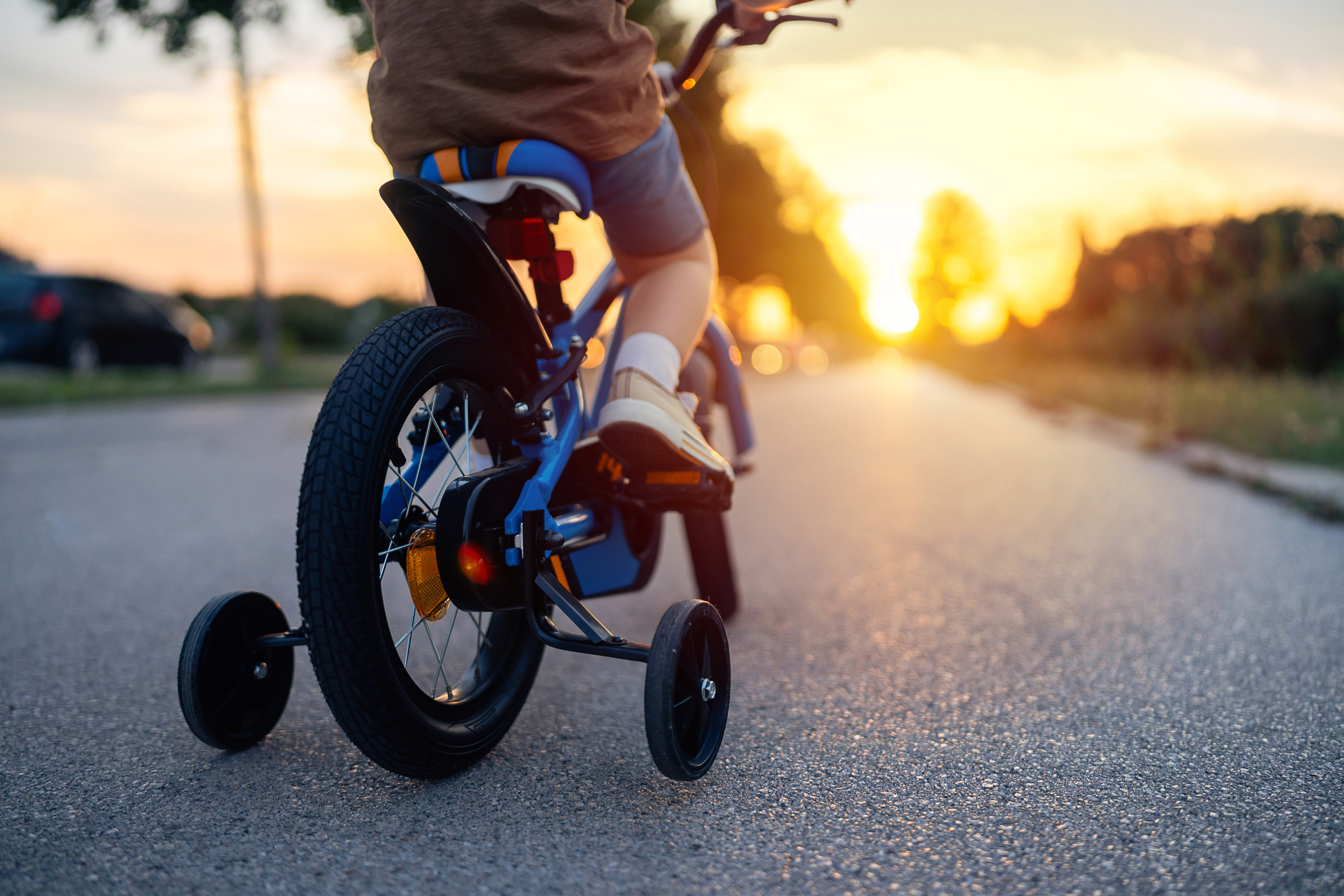 A child riding a bicycle with training wheels on a street at sunset, viewed from behind