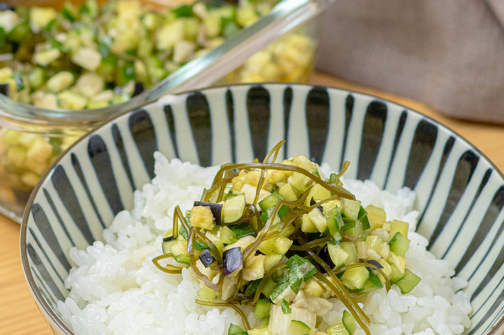 ストライプ模様の丼に盛り付けられた白米の上に、みじん切りの野菜と昆布がのっている料理。背景には調理中の姿。
