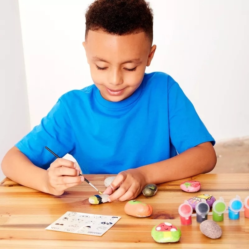 A child in a blue shirt sits at a wooden table painting small stones using a brush and several paint colors