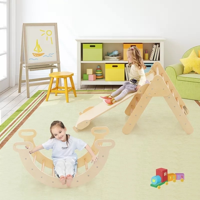 Two children are playing with wooden indoor climbing toys in a playroom. A shelf with toys and an easel with a boat drawing can be seen in the background