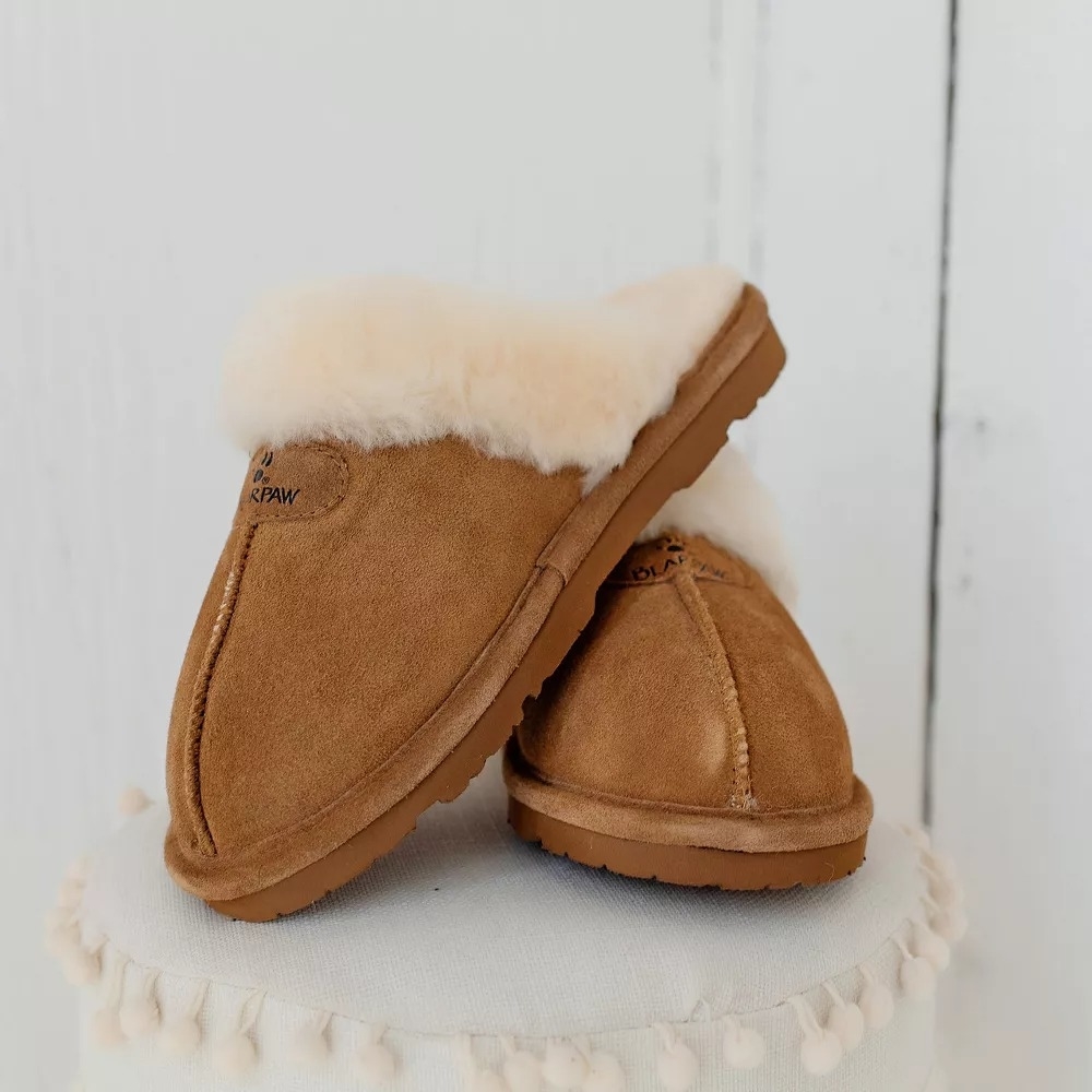 A pair of suede and shearling slippers, positioned with one standing upright and the other leaning against it, displayed on a white surface with pom-pom trim