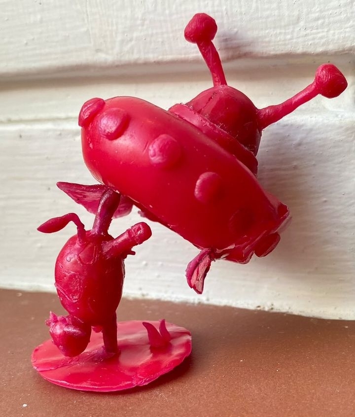 Red wax sculpture of two whimsical, ant-like creatures interacting, one appearing to carry the other