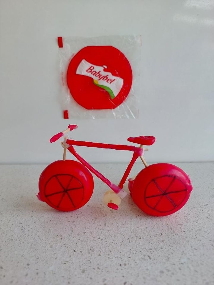 A miniature bicycle crafted from Babybel cheese wax, with an unwrapped Babybel cheese and its packaging in the background