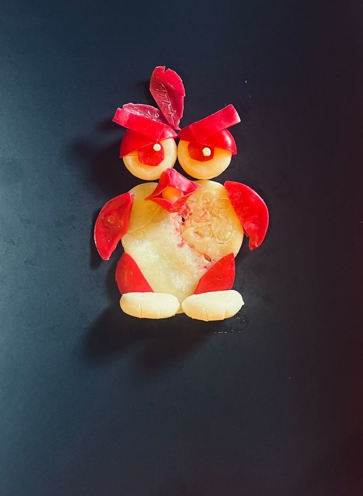 A fun arrangement of sliced fruits and vegetables resembling an angry bird character with expressive eyes and red feathers