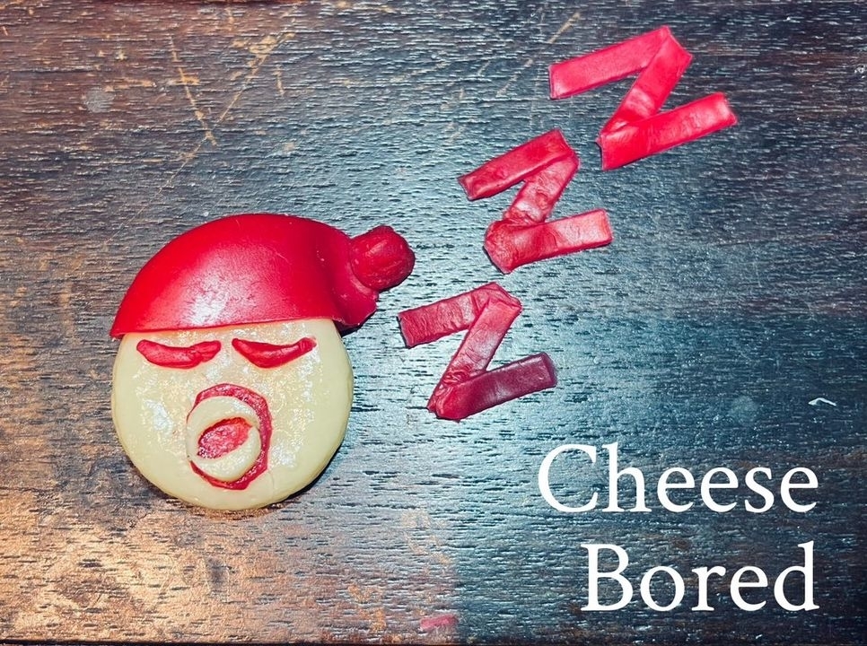 Round piece of cheese with a red cap and facial features made from red vegetable slices, surrounded by &quot;Zzz&quot; letters and the text &quot;Cheese Bored&quot; on a wooden surface