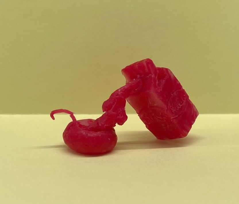 A small, intricately carved red wax sculpture, featuring abstract shapes and objects, is displayed against a plain background