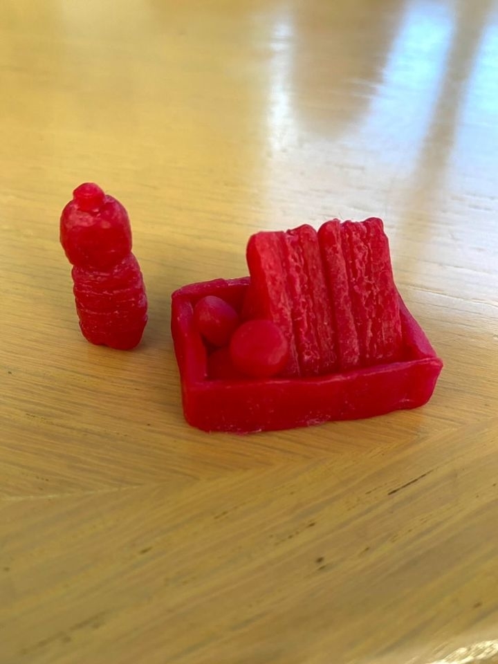 Red gummy candy shaped like a bottle, two balls, and a tray with long rectangular pieces resembling bacon strips on a wooden table