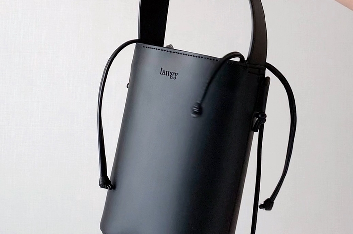 A person’s hand, wearing a wristwatch, holds a stylish, cylindrical black handbag with a handle and drawstring closure. The word “lawgy” is on the bag