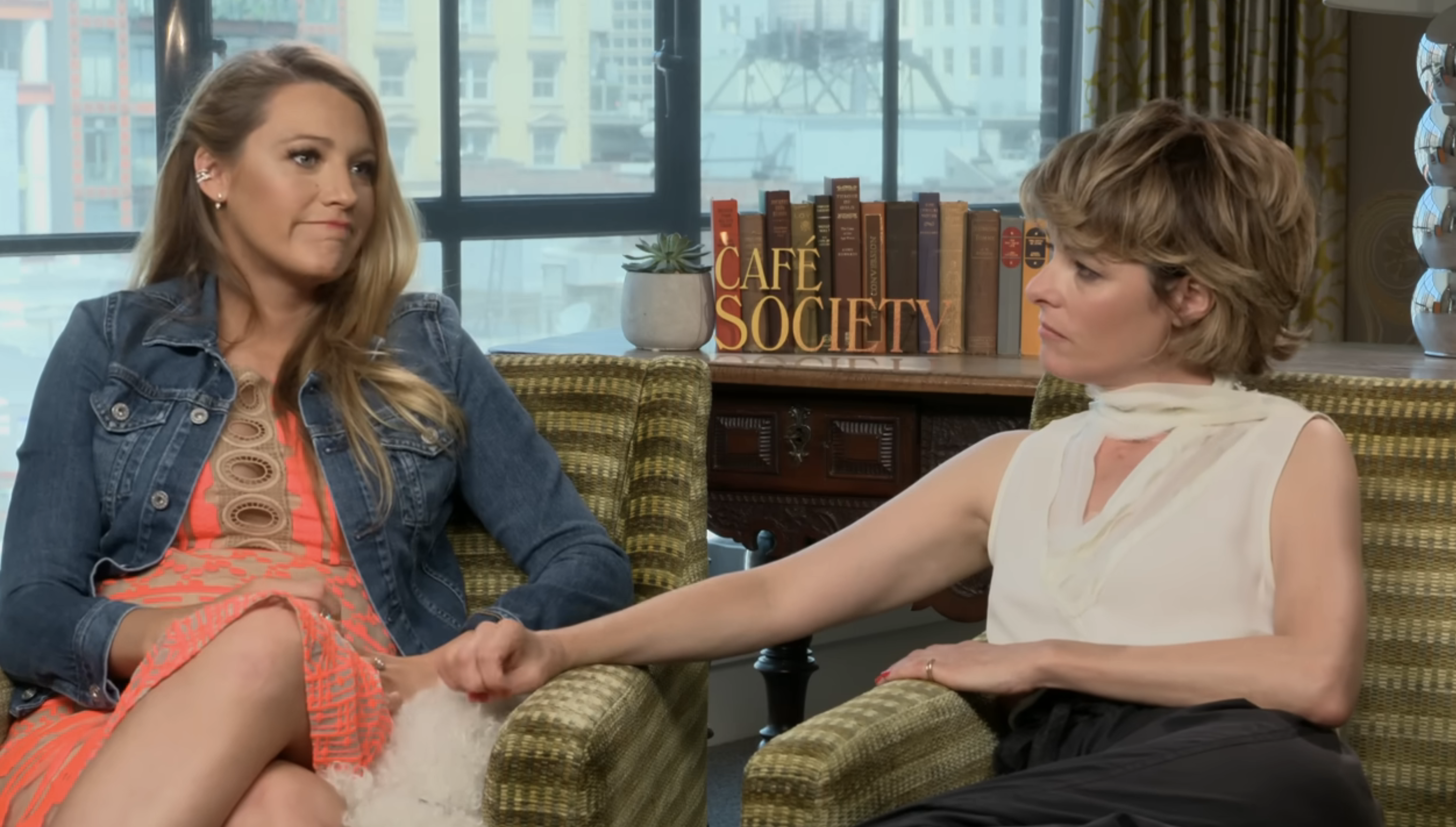 Blake Lively in a denim jacket and Parker Posey in a sleeveless white blouse sit on a sofa holding hands, with a &quot;Café Society&quot; sign in the background