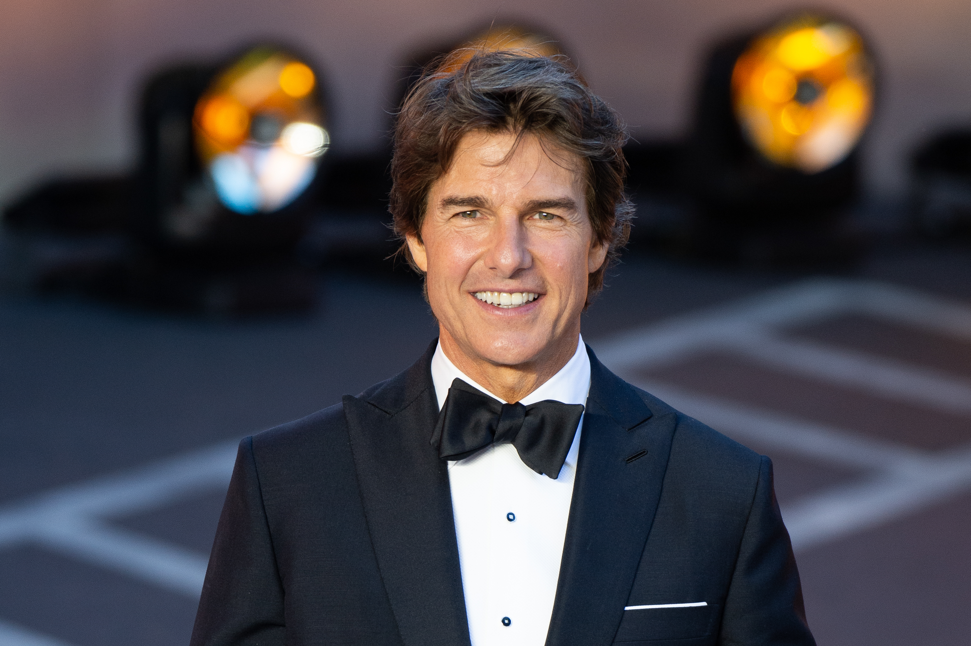 Tom Cruise is on the red carpet in a black tuxedo with a bow tie, smiling for the camera