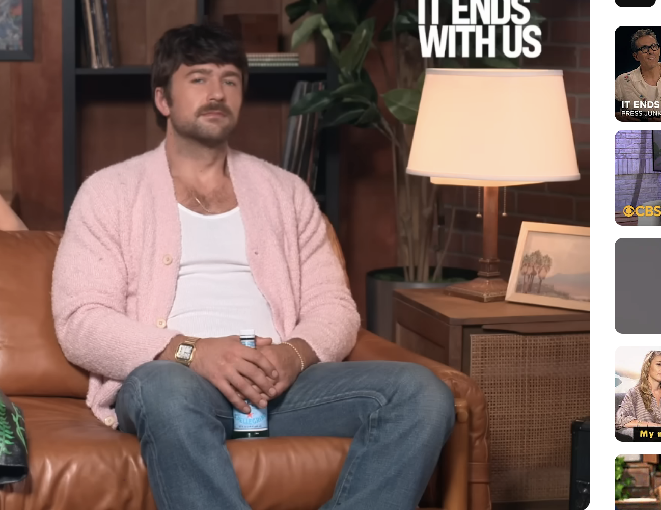 Blake Lively, in a printed dress, and Brandon Sklenar, in a white shirt and pink cardigan, sit on a couch during an interview. &quot;IT ENDS WITH US&quot; text in the background