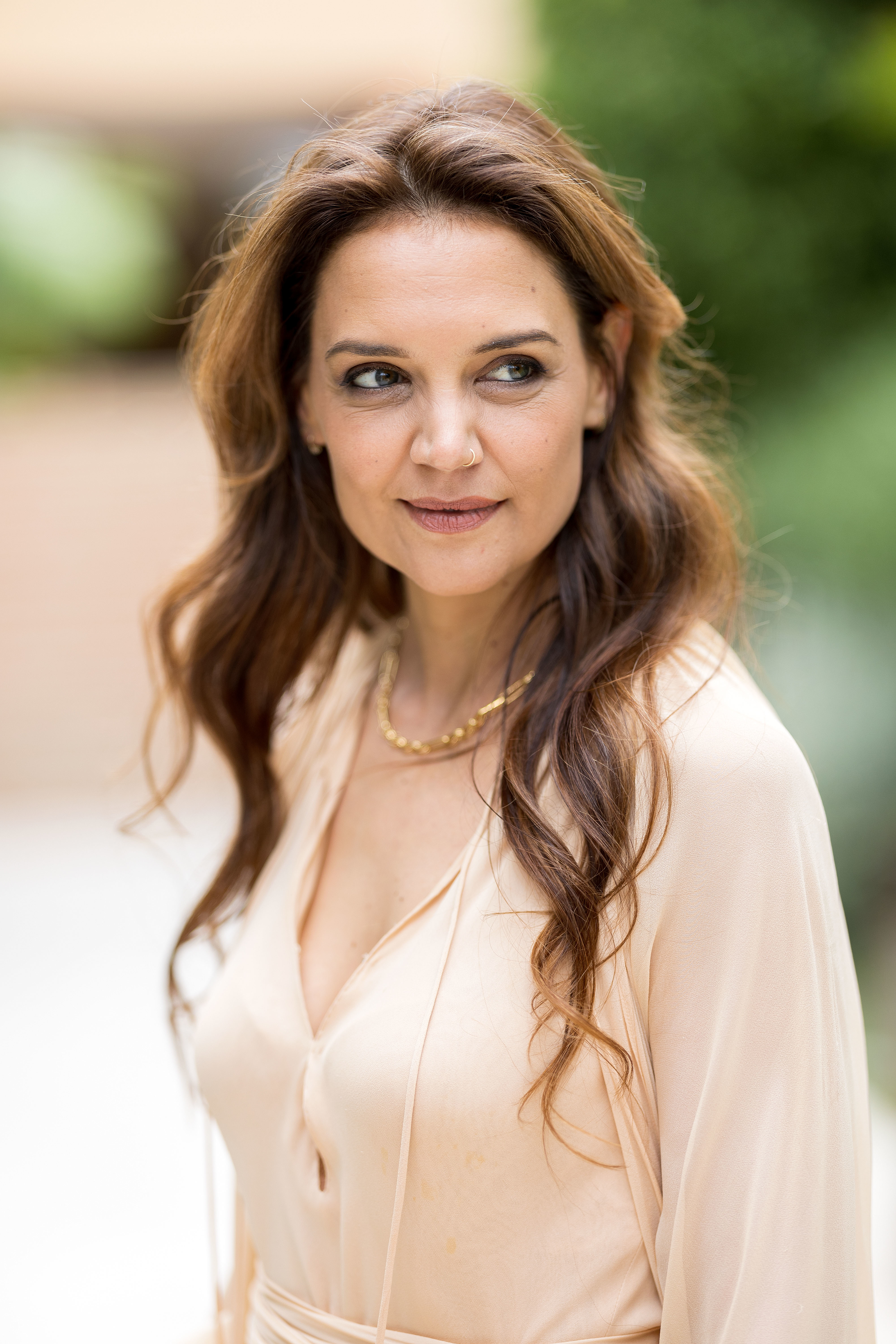 Katie Holmes wearing a flowing V-neck blouse, standing outdoors, with loose wavy hair, a simple gold necklace, and a slight smile