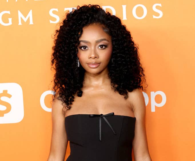 Skai stands in front of an backdrop at an event, wearing a strapless dress with a small bow