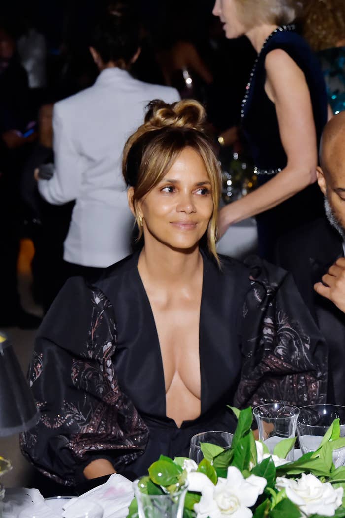 Halle Berry sits at an event table, wearing a deep V-neck gown with puffy sleeves. Other guests are visible in the background