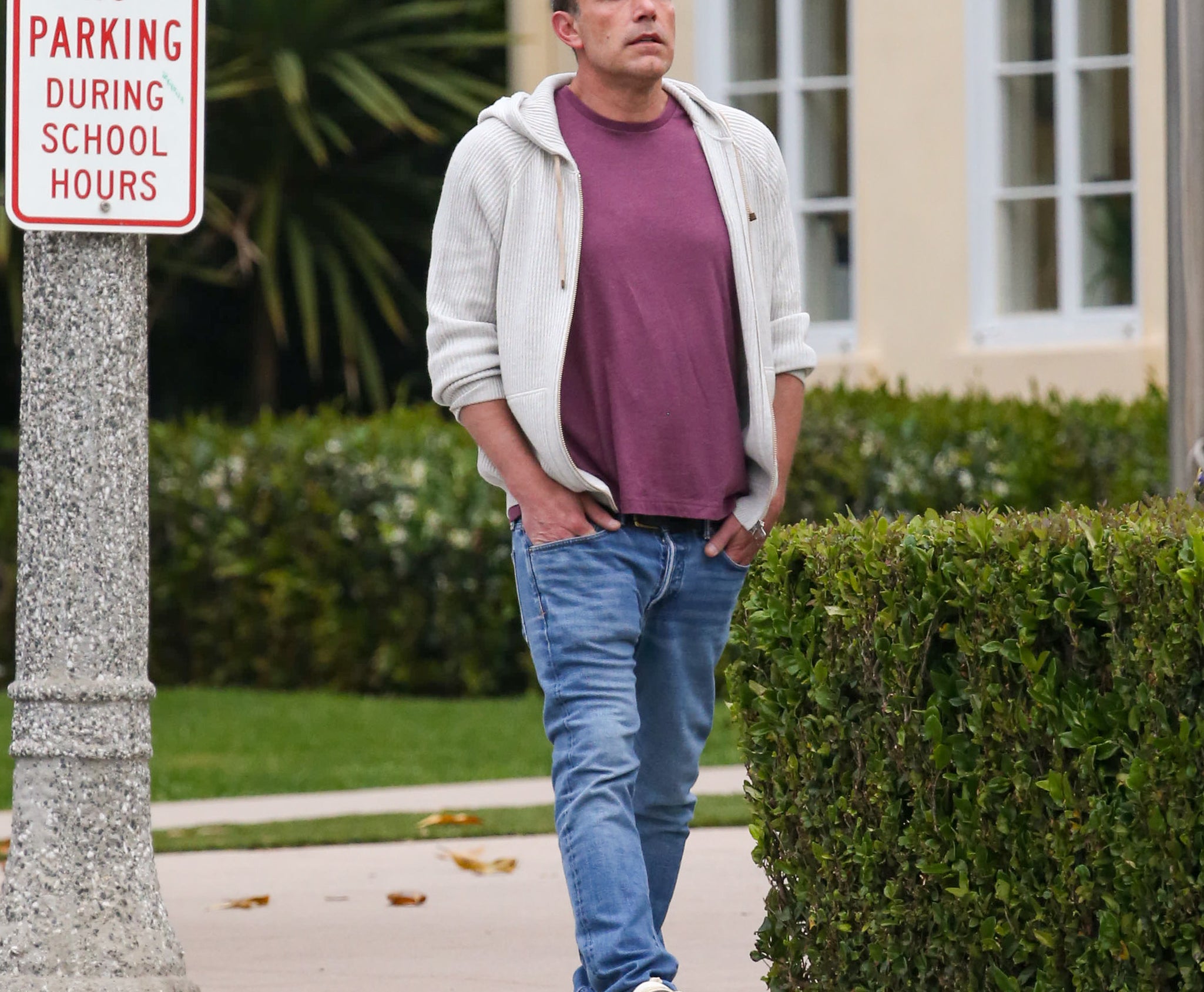 Ben Affleck in casual attire, wearing a purple shirt, jeans, and a white hoodie, walks on a sidewalk beside a &quot;No Parking During School Hours&quot; sign