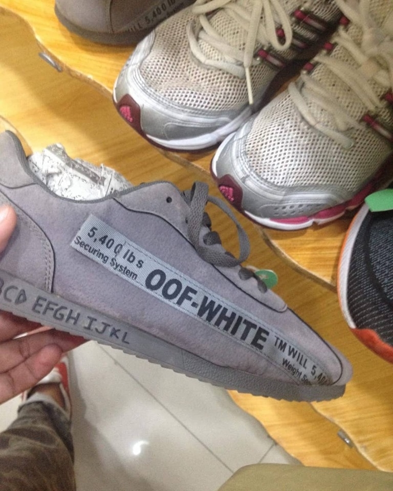 A hand is holding a gray sneaker labeled with "OOF-WHITE" and various text details. Other pairs of sneakers are visible in the background