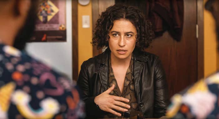 Ilana Glazer, with curly hair, sits at a table gesturing, wearing a leather jacket, talking to two unseen people