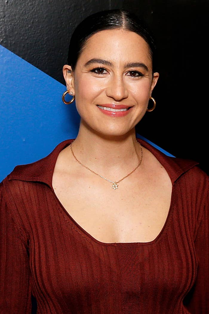 Ilana Glazer smiles, wearing a ribbed top with a low neckline, a star-shaped necklace, and hoop earrings