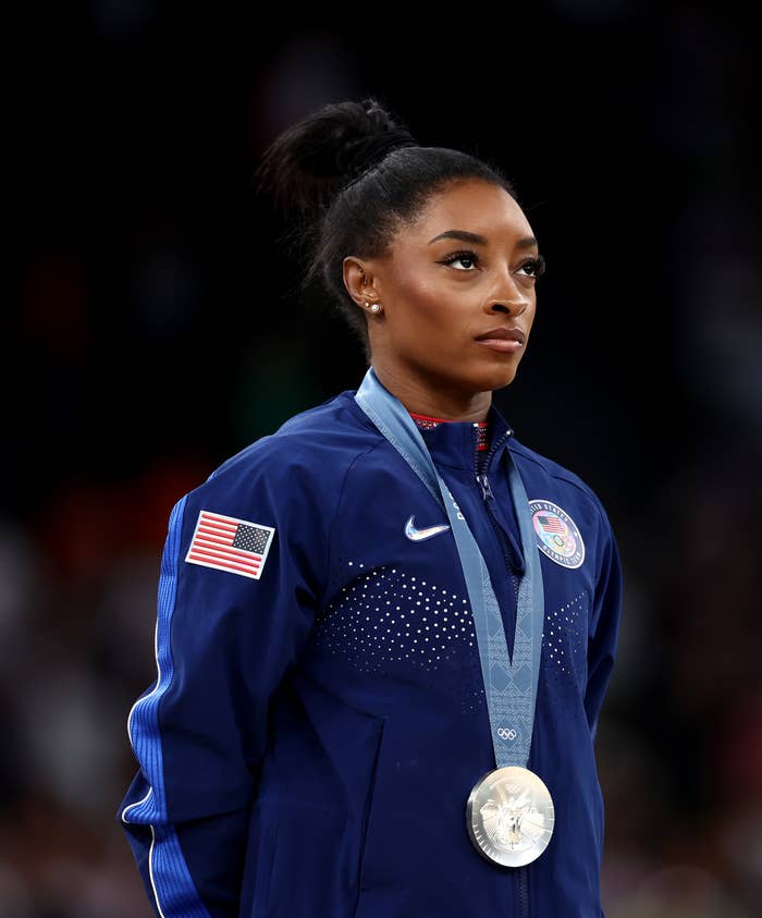 Simone Biles stands with a serious expression, wearing a USA track jacket and a silver medal around her neck