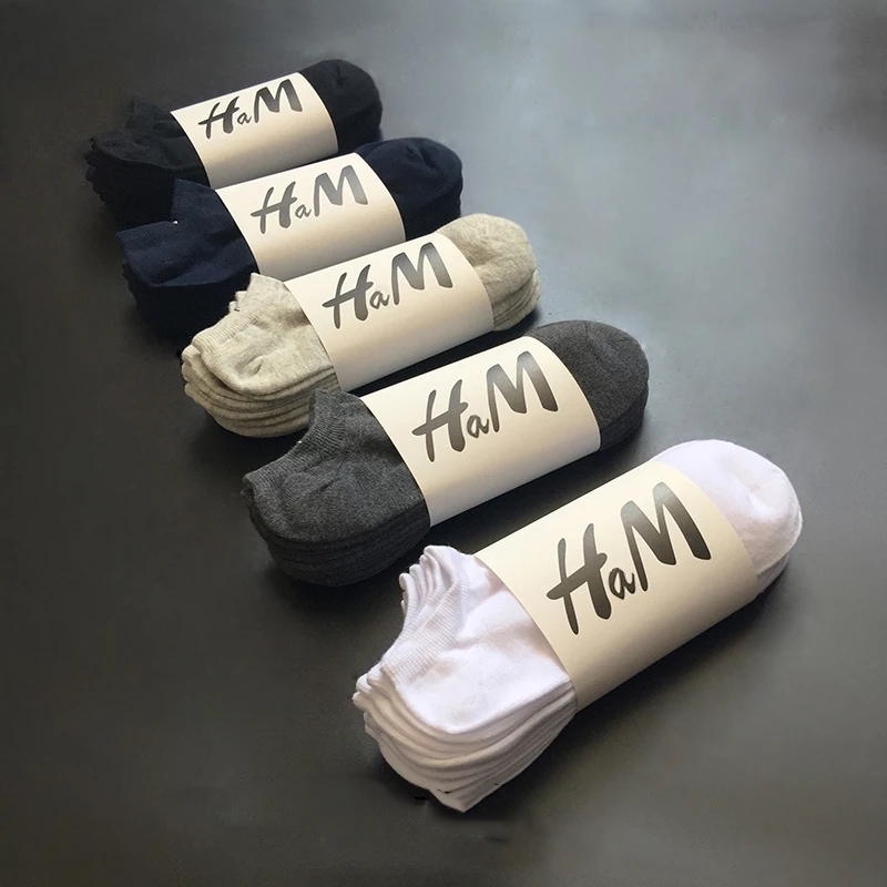 Five pairs of socks, each wrapped in HaM branded packaging, are displayed in a row on a dark surface. The colors include black, navy, grey, dark grey, and white