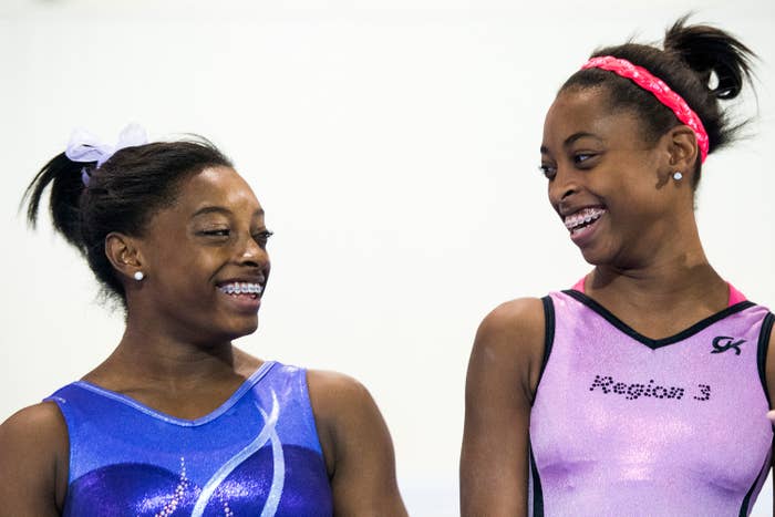 A younger Simone Biles and another gymnast smiling at each other, wearing sleeveless gymnastics leotards