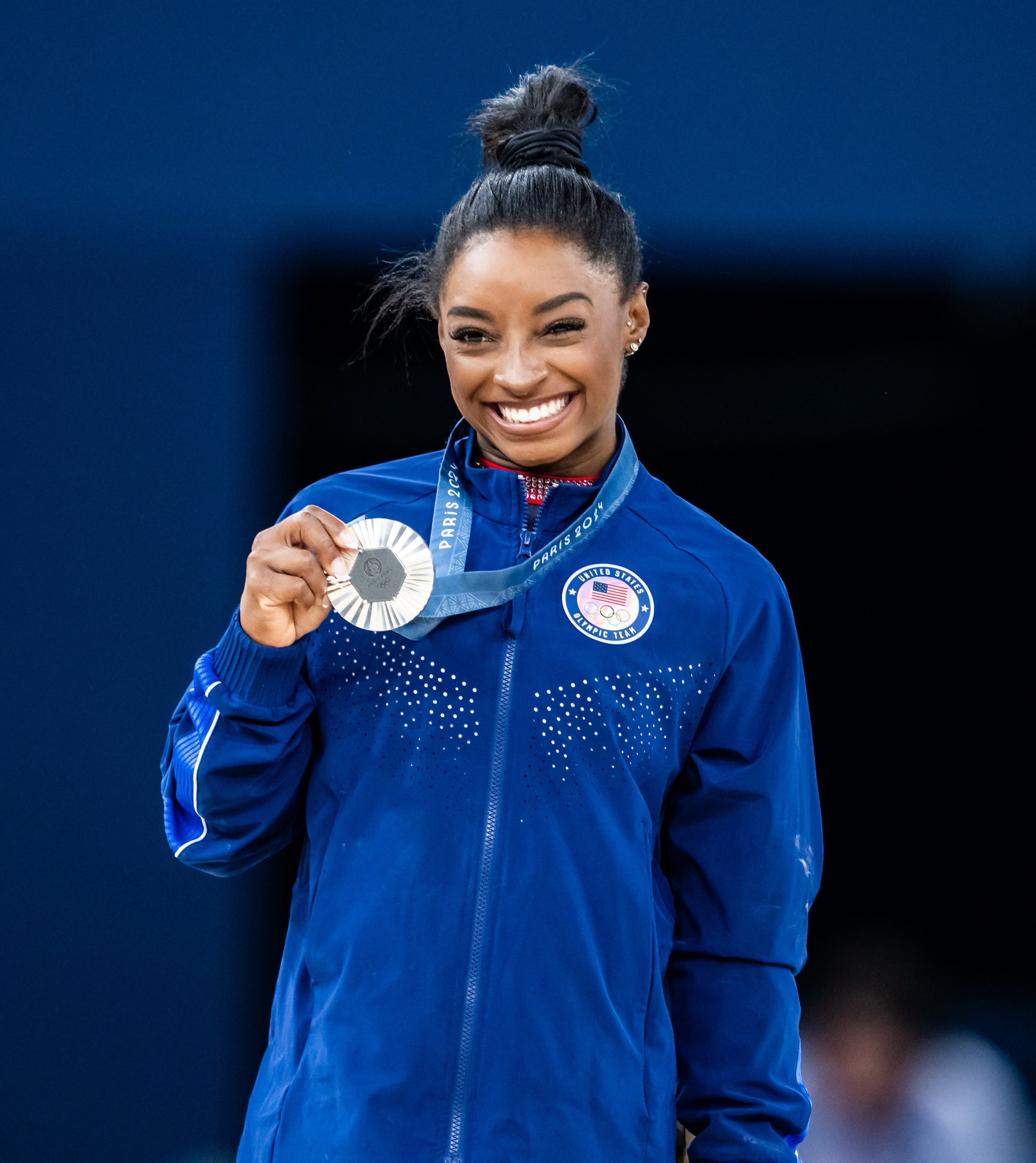 Simone Biles smiles and holds a silver medal while wearing a tracksuit