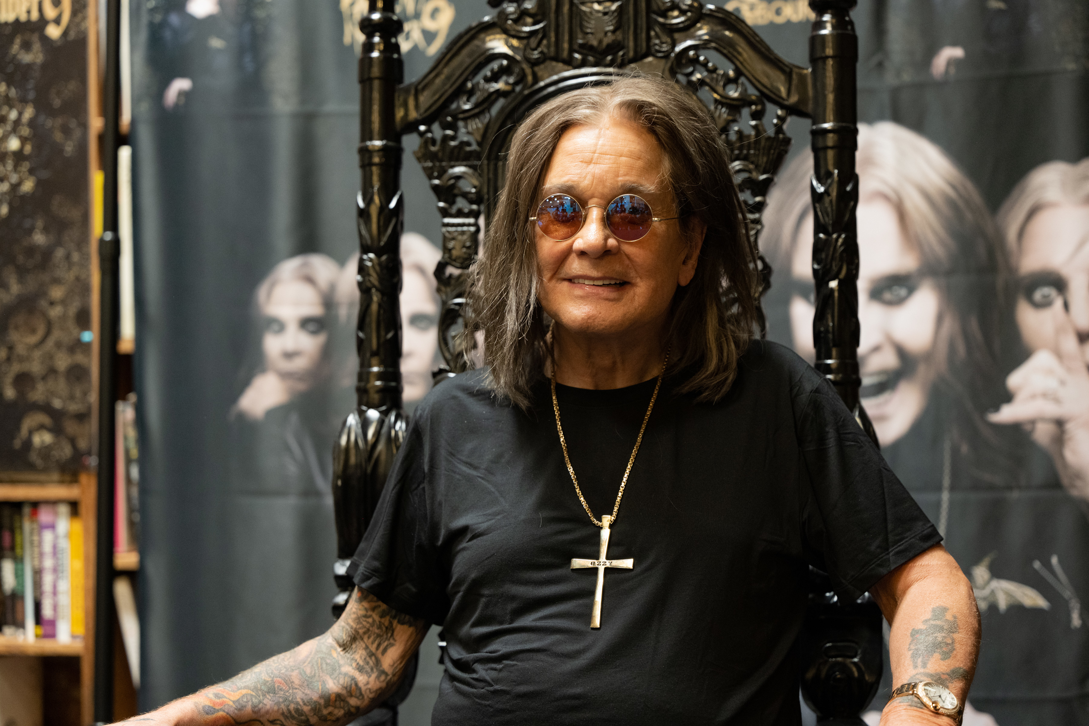 Ozzy Osbourne sits on a throne-like chair wearing round sunglasses and a t-shirt with a cross necklace. Posters with his image are in the background