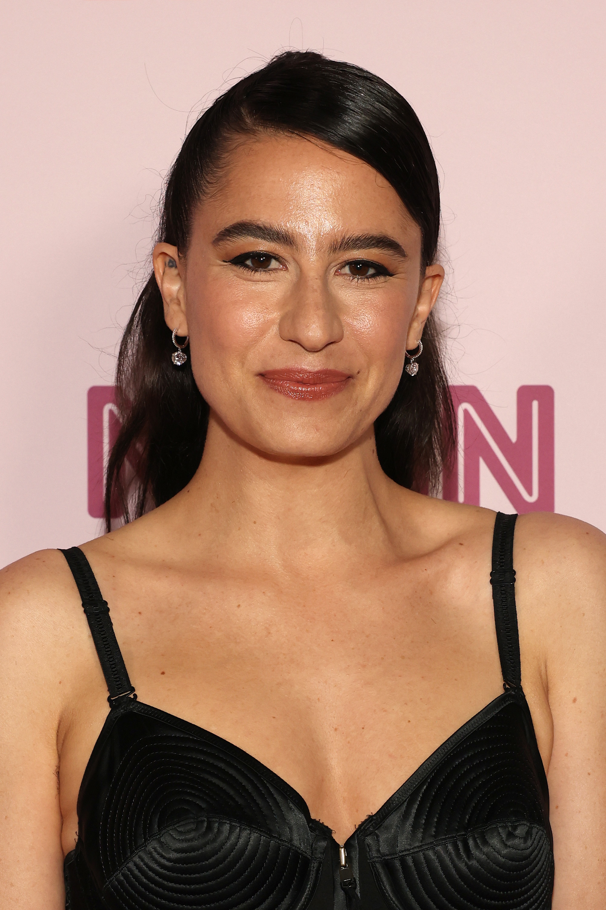 Ilana Glazer, smiling, poses in a black sleeveless dress with geometric patterns detail on the red carpet