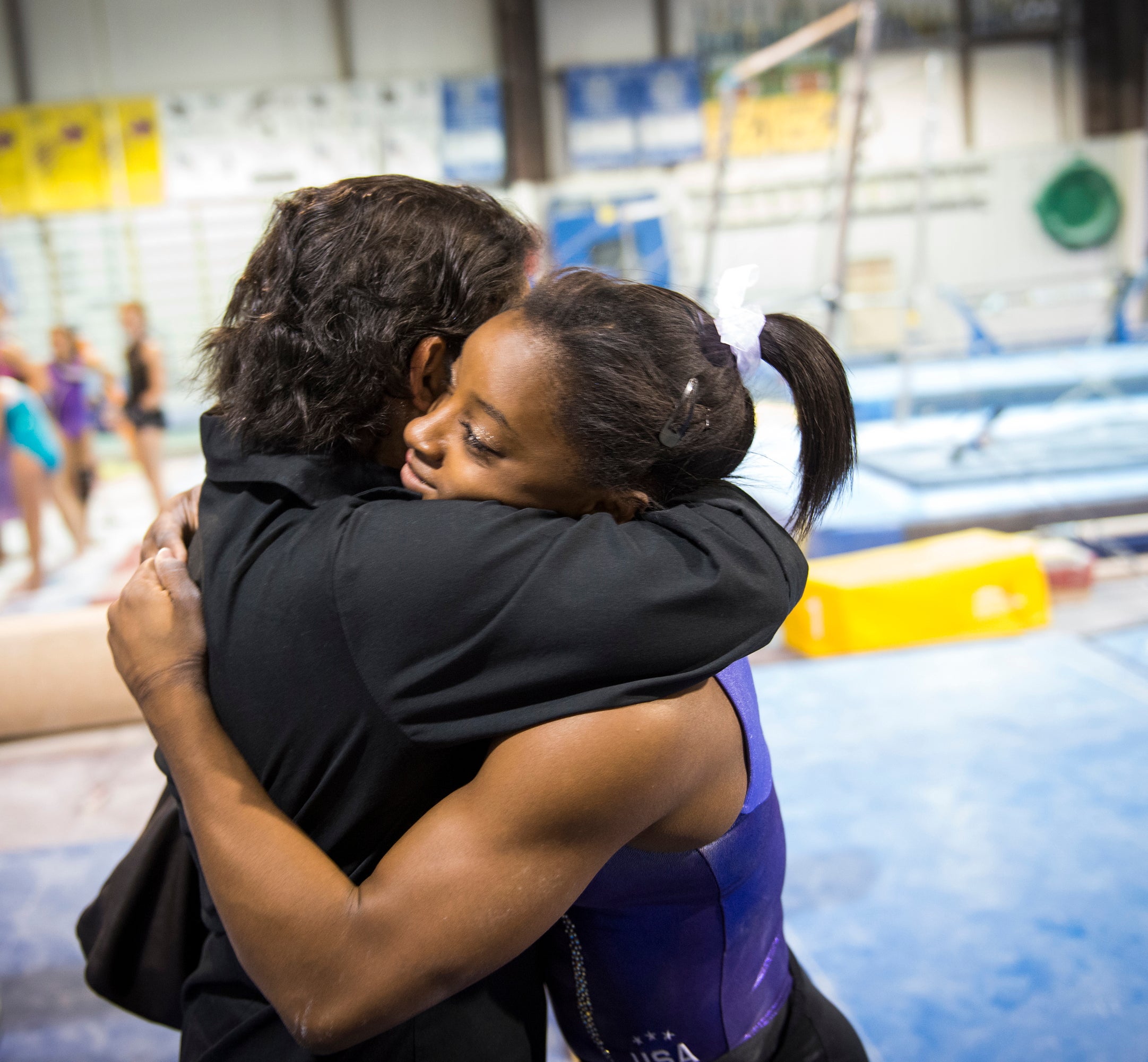 Simone Biles hugs her mom, Nellie, in a gymnastics training facility. Gymnastics equipment and other athletes are visible in the background