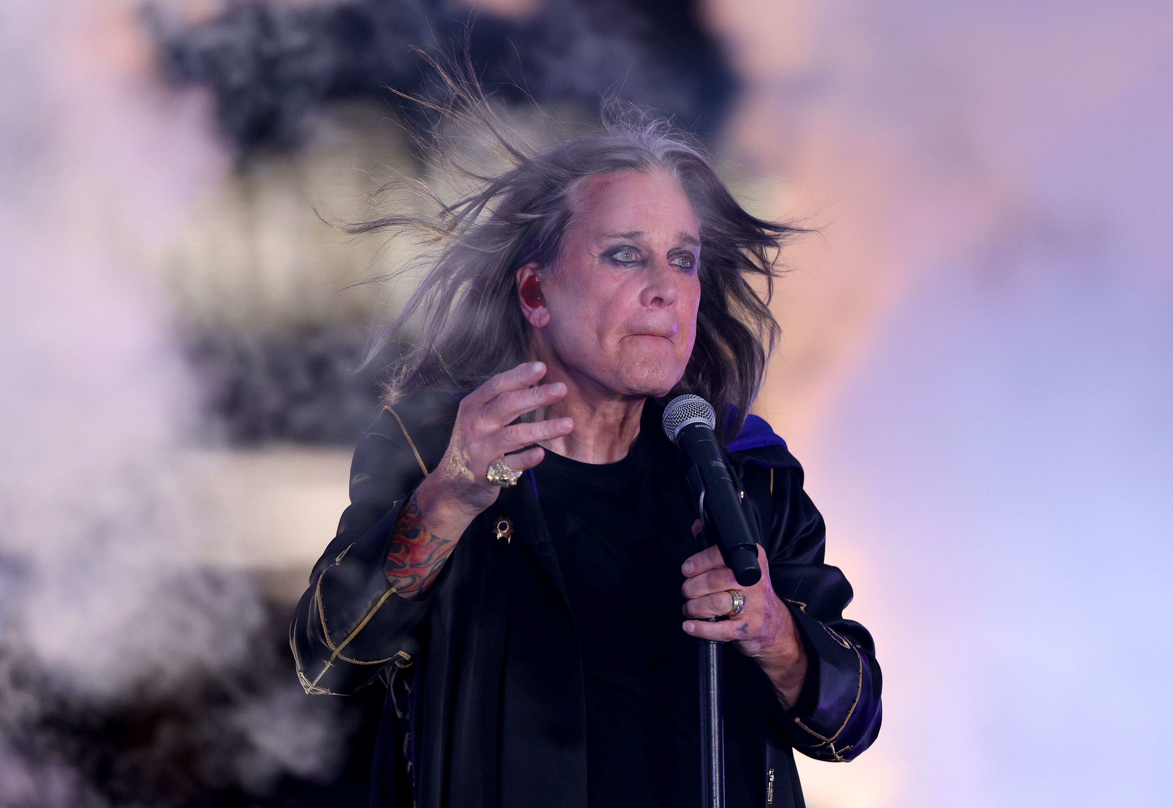Ozzy Osbourne performs on stage, holding a microphone. His hair flows in the wind as he wears a dark outfit with a coat