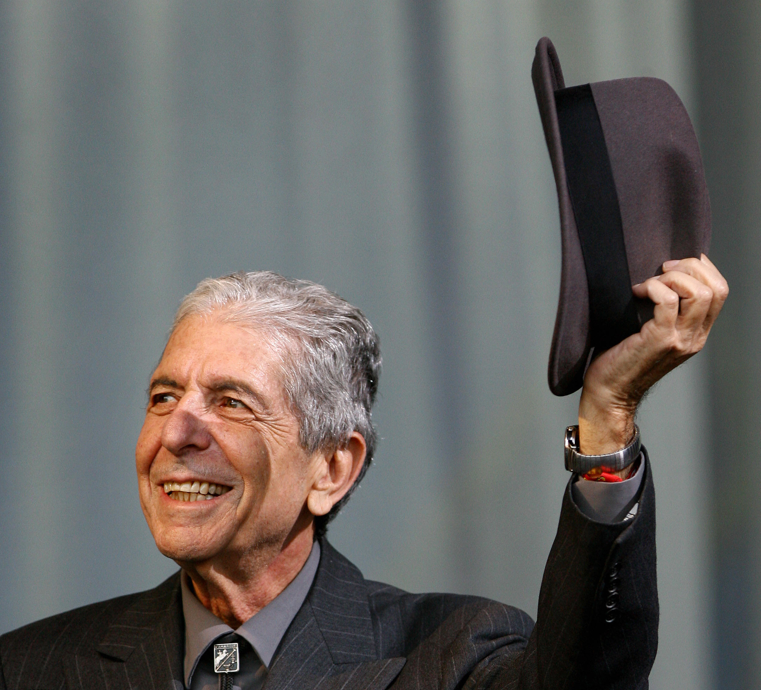 Leonard Cohen smiling, holding a hat in his right hand, wearing a pinstripe suit and tie