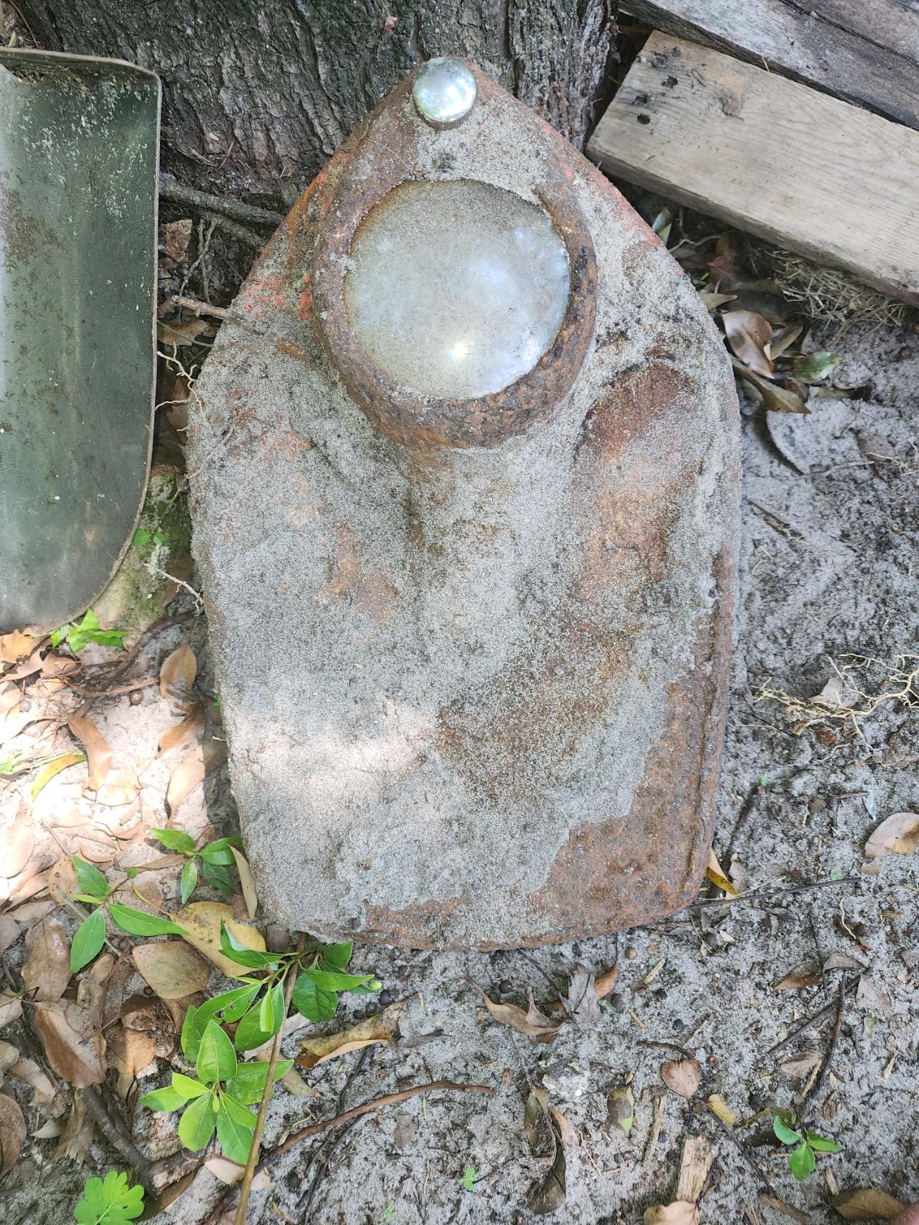 Rusty metal object partially buried in dirt, appearing to be an old, weathered iron with a large round glass panel on top. Background features tree roots and wooden planks