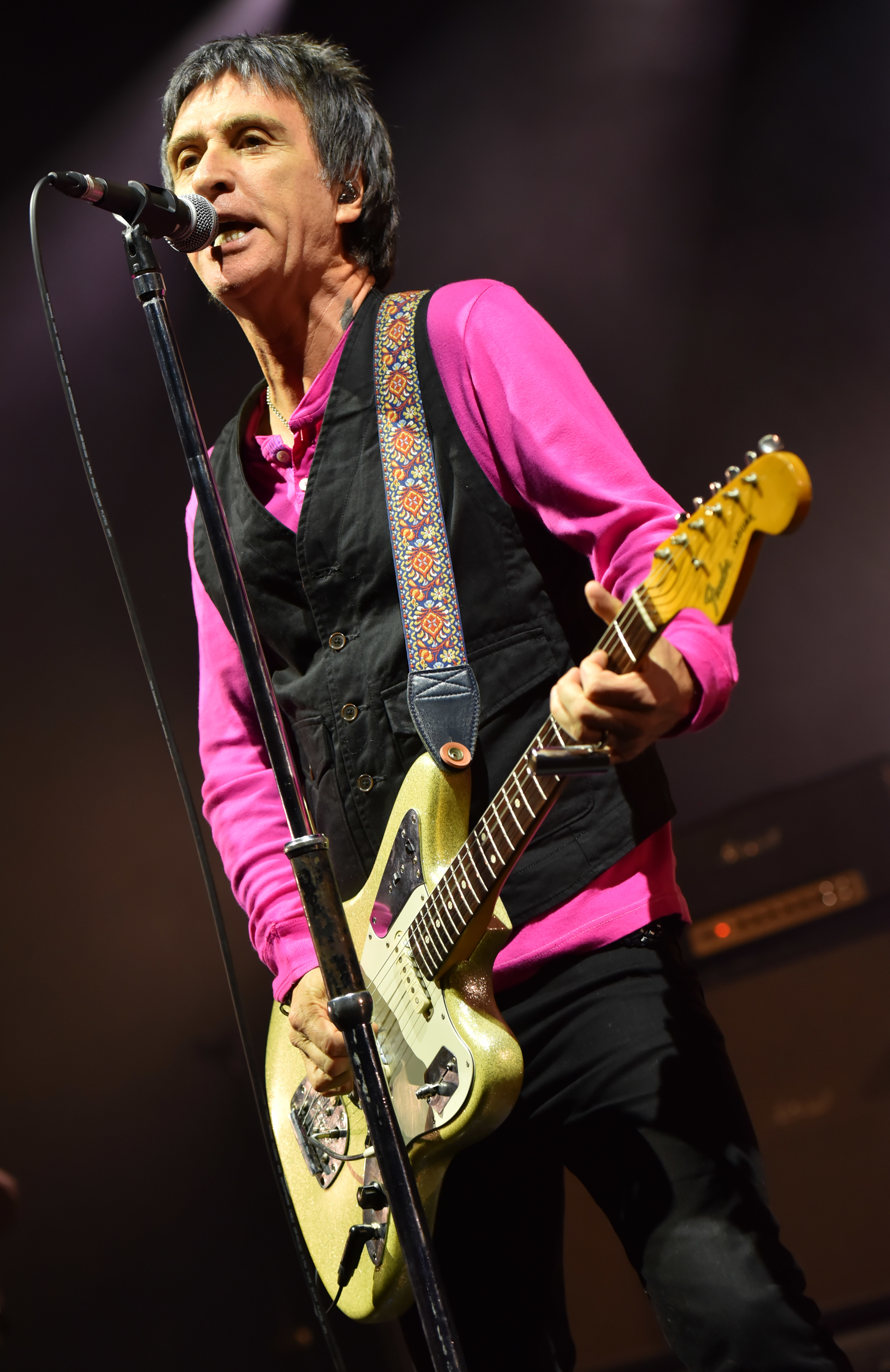 Johnny Marr performs on stage, wearing a black vest over a pink shirt, playing a Fender guitar and singing into a microphone