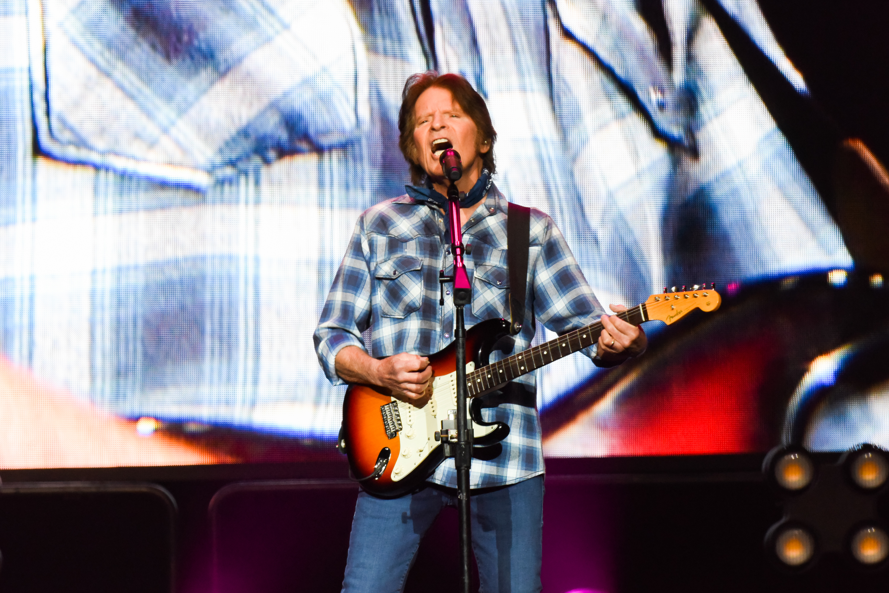 John Fogerty performing on stage, wearing a plaid shirt and jeans, playing an electric guitar, and singing into a microphone. Large screen backdrop behind him