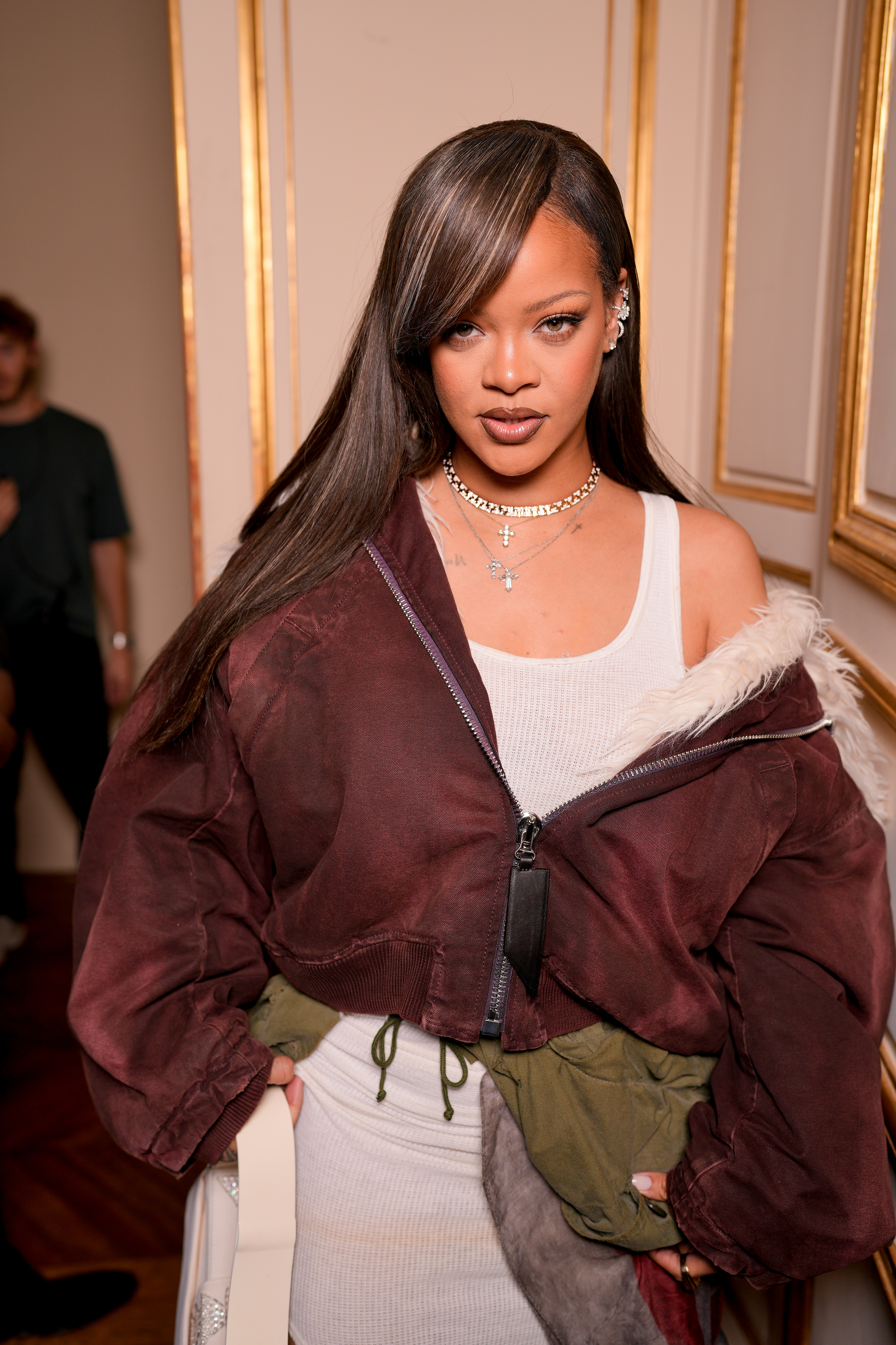 Rihanna poses indoors wearing a stylish jacket over a dress. She sports layered necklaces and side-swept hair