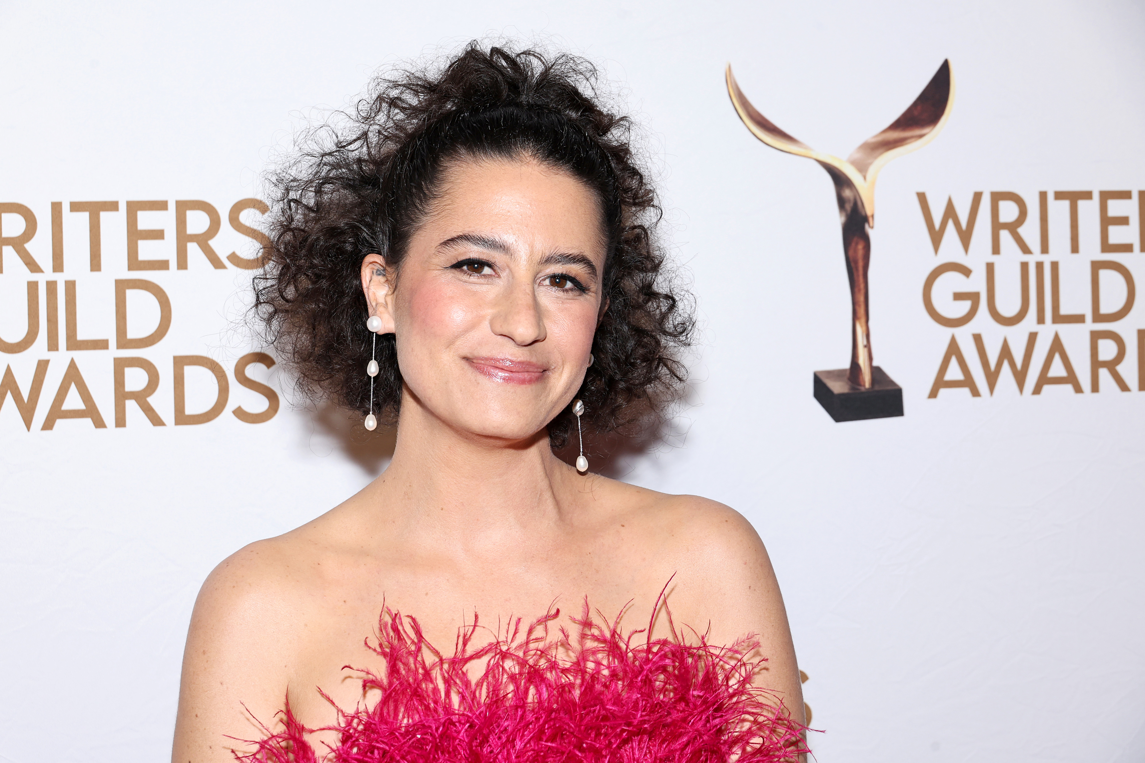 Ilana Glazer stands smiling at the Writers Guild Awards. She wears a pink dress with feathery embellishments and drop earrings