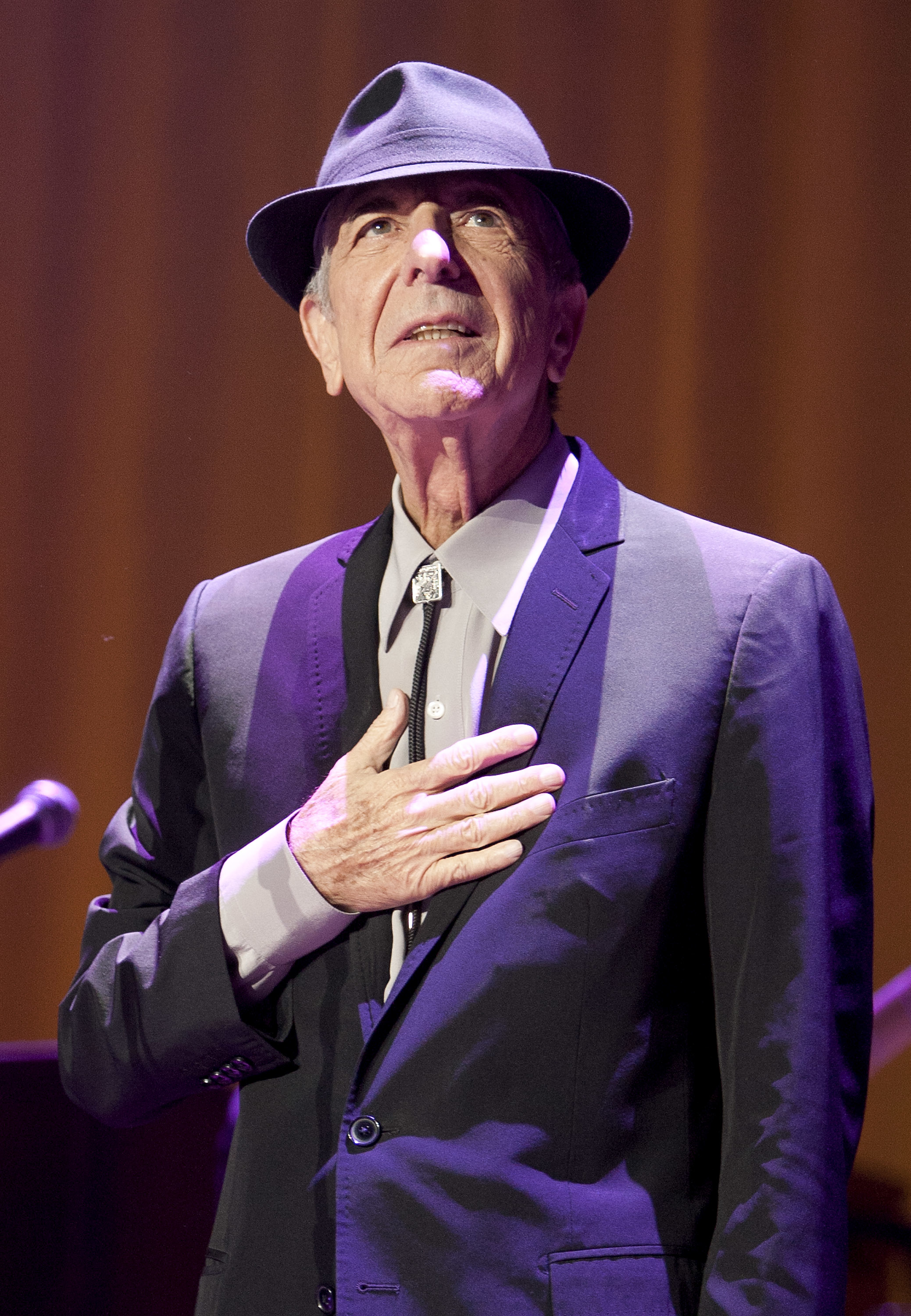 Leonard Cohen, dressed in a dark suit and hat, stands with his hand over his heart during a performance