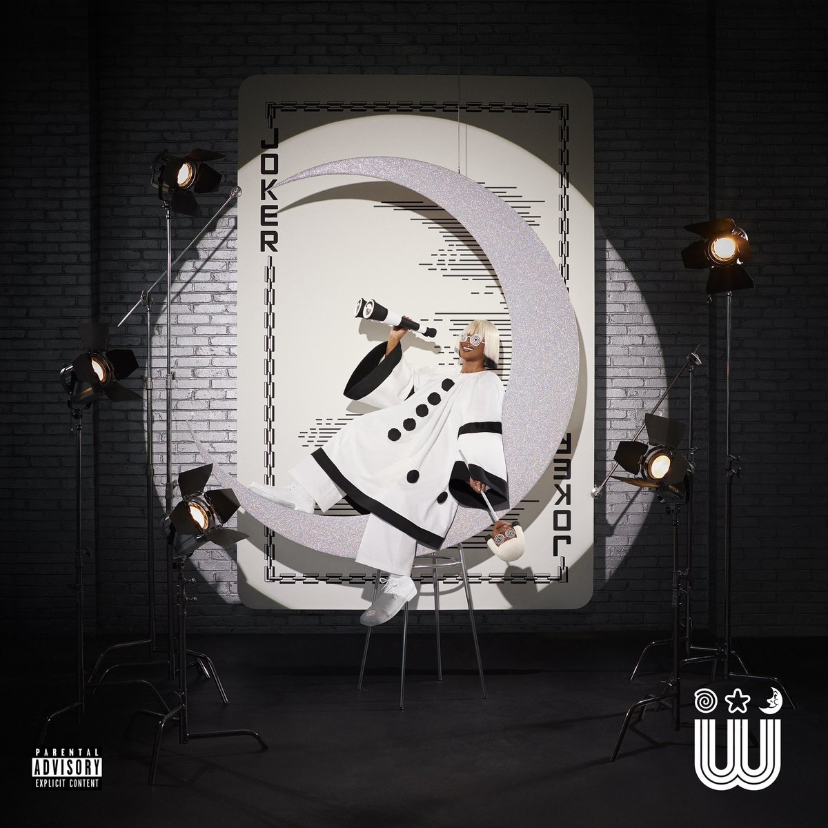 Tierra Whack poses in a theatrical costume resembling a clown, seated on a crescent moon in front of a large playing card backdrop. The image includes an explicit content advisory