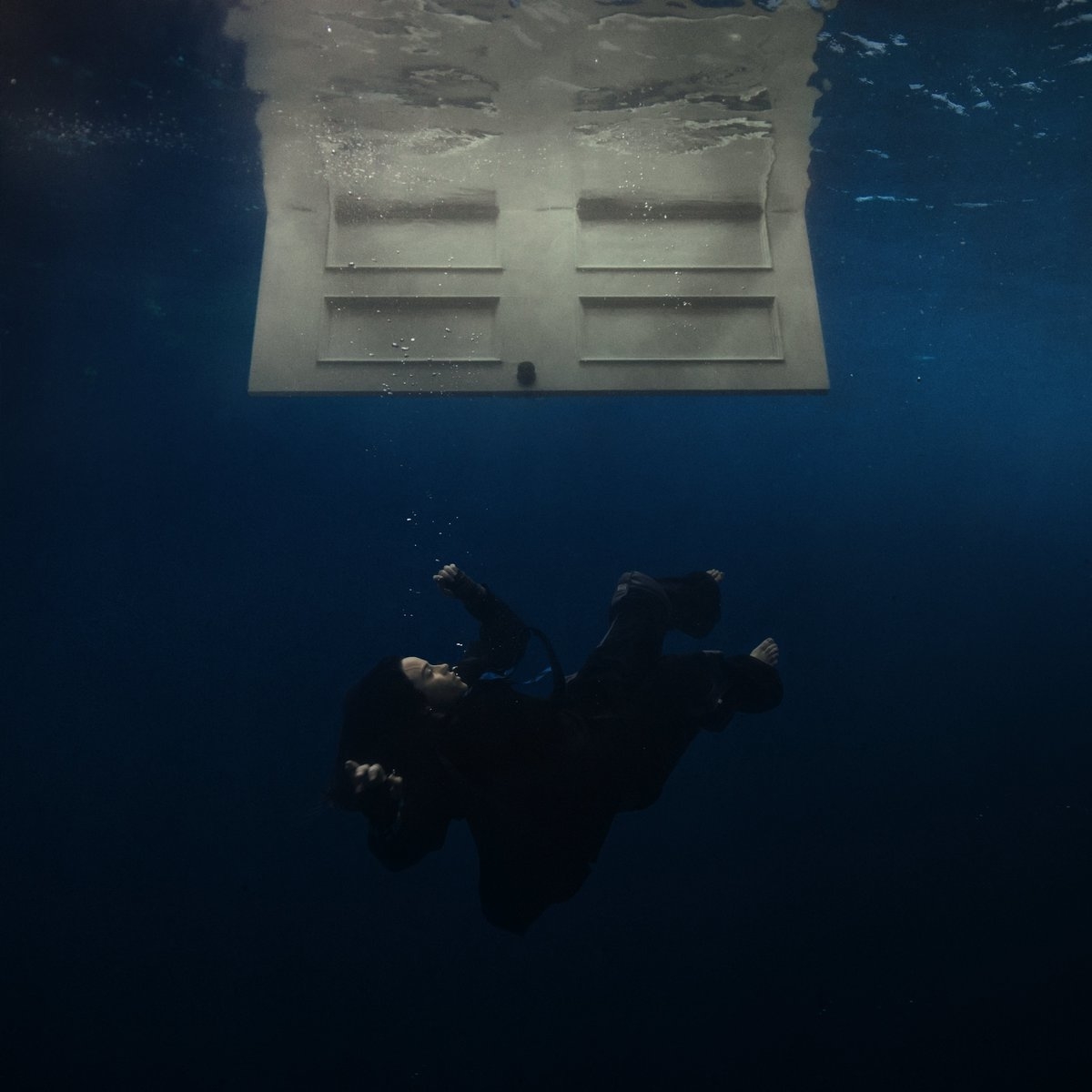 Billie Eilish in dark clothing floating underwater near an upside-down door