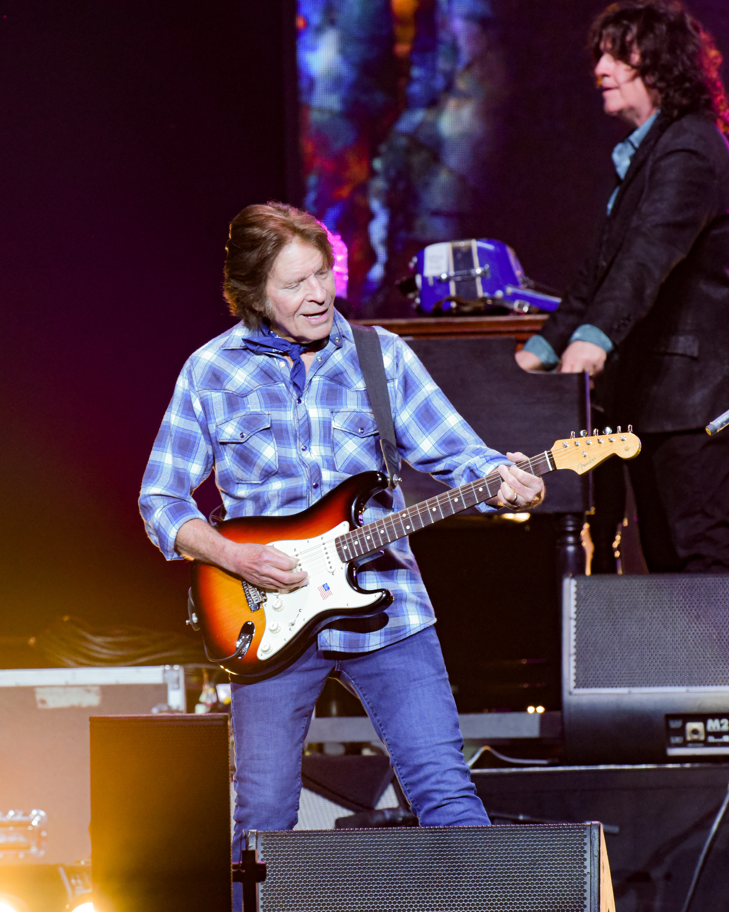 John Fogerty playing an electric guitar on stage, dressed in a casual plaid shirt and jeans, with a band member in the background