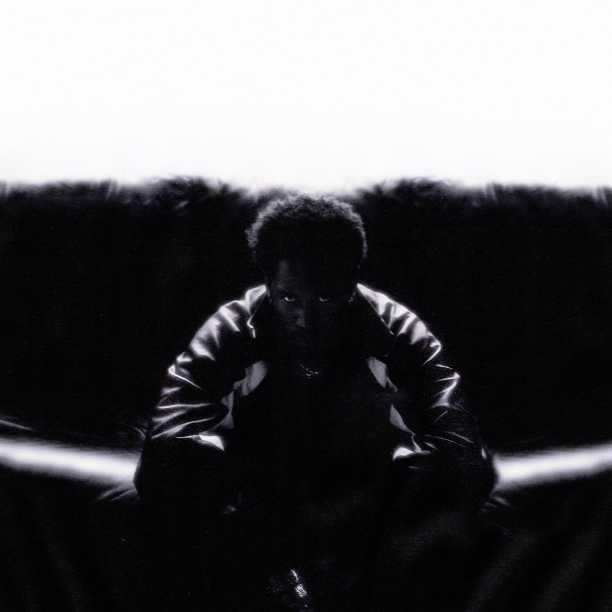 Kaytranada sits with a serious expression on a dark couch, wearing a jacket. The image conveys a moody and intense ambiance