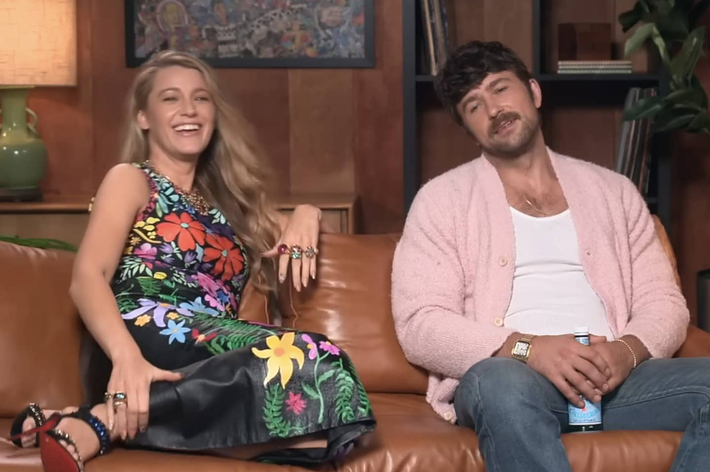 Blake Lively, wearing a floral dress, laughs while sitting on a sofa beside Brandon Sklenar, dressed casually in a pink cardigan and white shirt