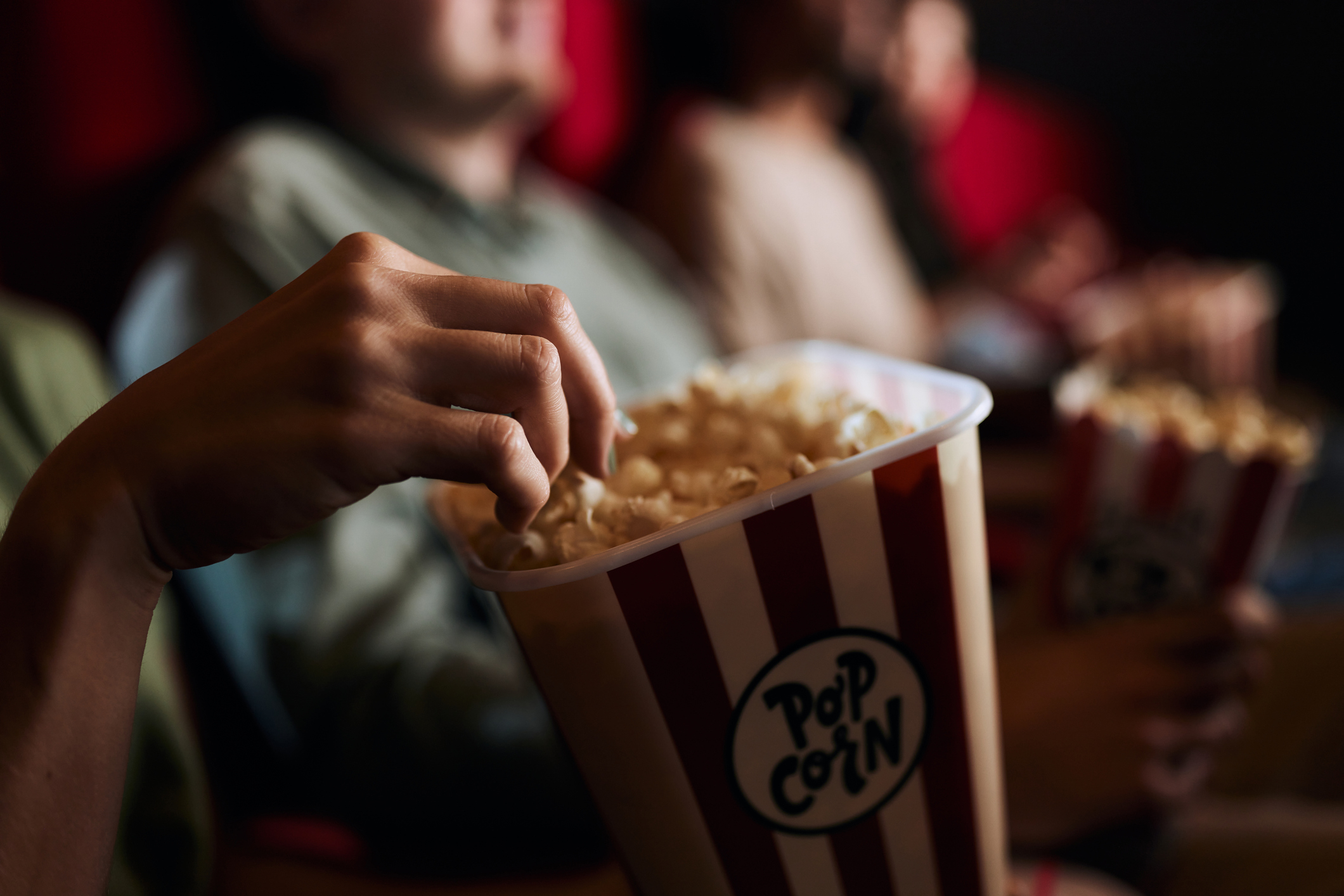 People in a cinema, focusing on a hand reaching into a striped popcorn tub labeled "Popcorn."