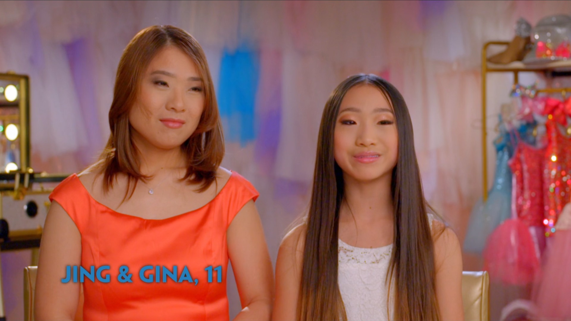 Jing and Gina are sitting together in a room with dresses in the background.