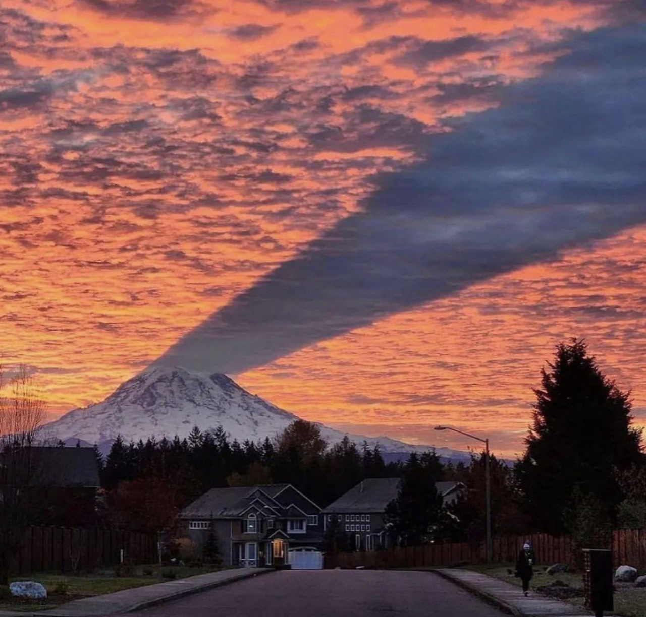 A picturesque sunset scene with a mountain casting a long shadow across the sky, viewed from a suburban street with houses, trees, and a person on a bike