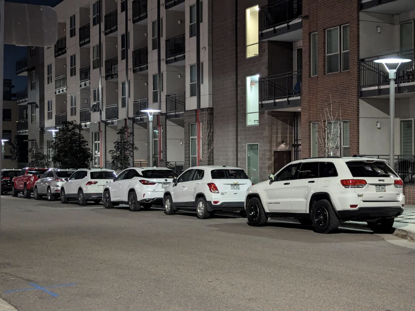 Several white SUVs and a few red cars are parked in a line along a residential street at night. Apartment buildings with balconies are visible in the background