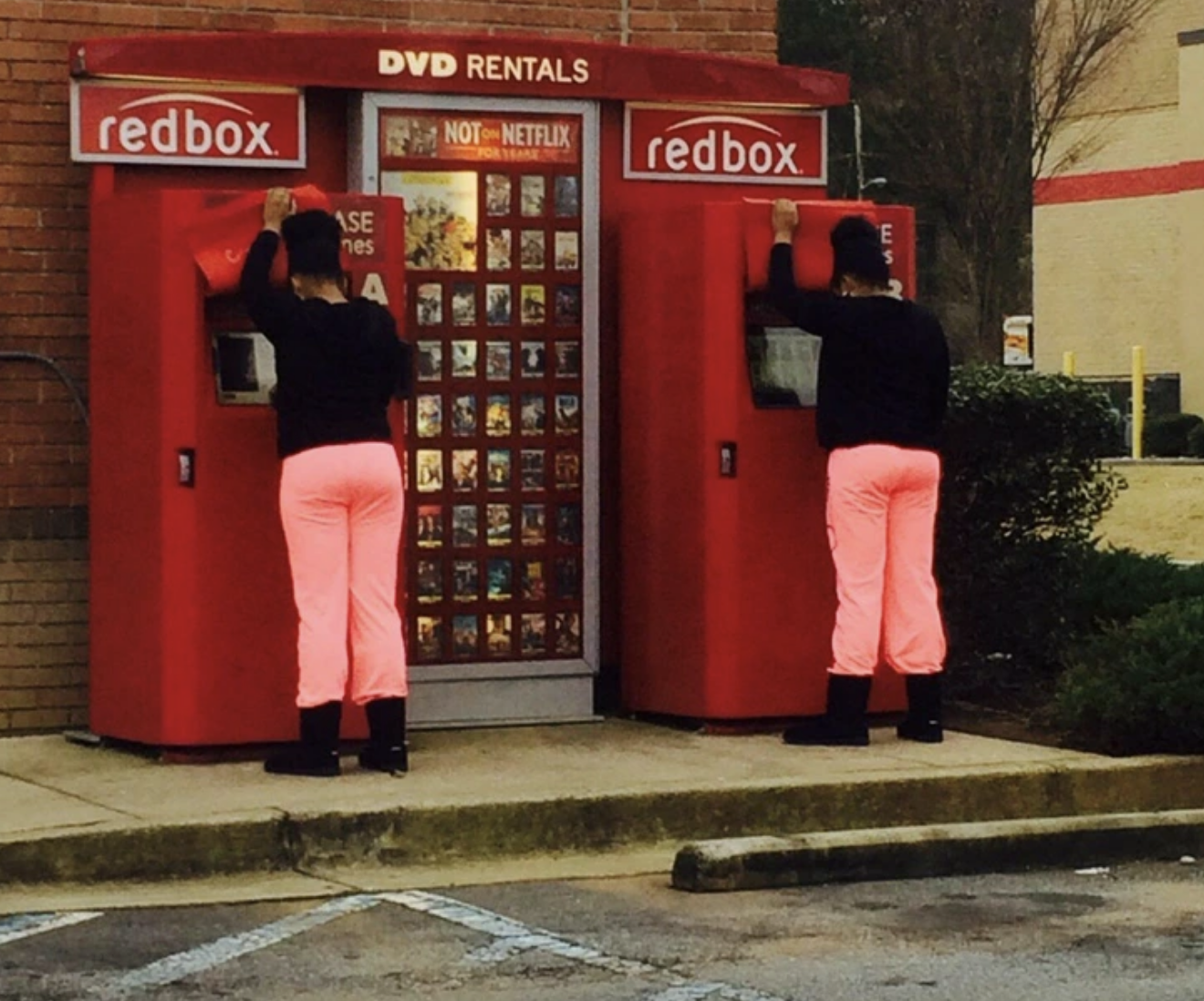 Two people are at Redbox DVD rental kiosks, both wearing black tops and pink pants