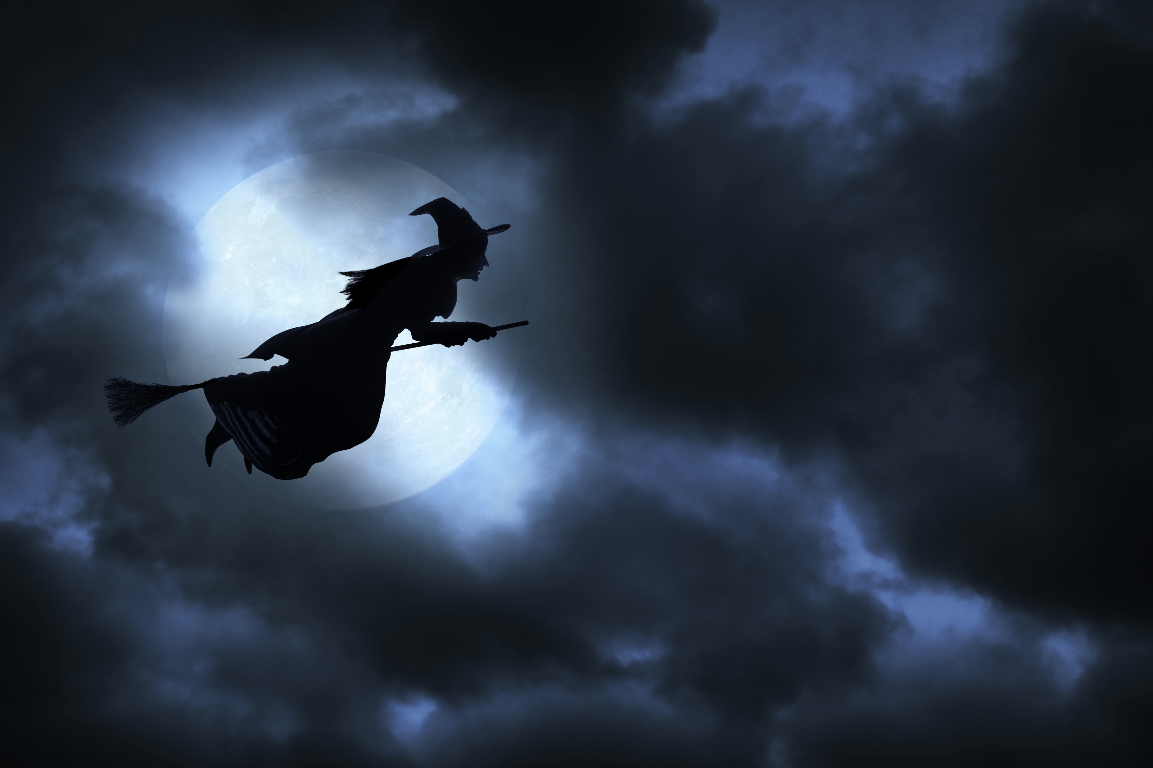 A silhouetted witch flies on a broomstick across a full moon, surrounded by clouds in a dark sky