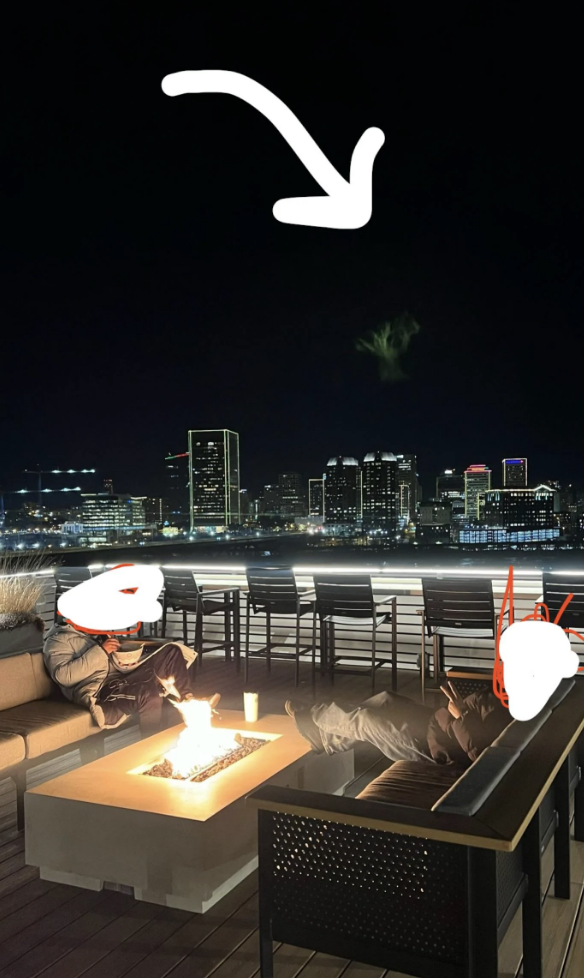Two individuals sit with drinks by a rooftop fire pit, enjoying a nighttime city skyline view. An arrow points to the sky, indicating something